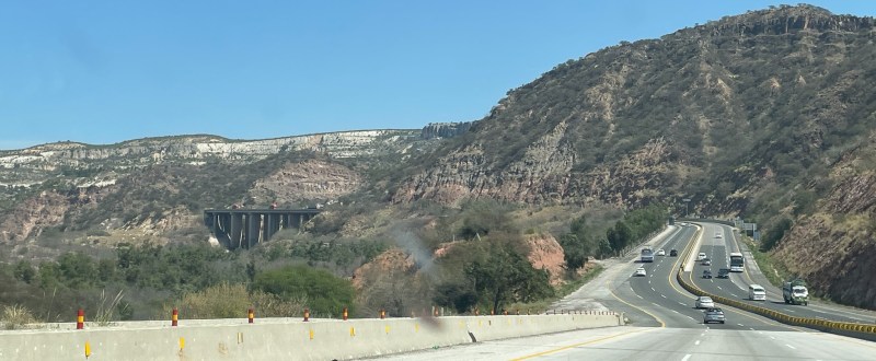View of the six lane M2 Lahore-Peshawar motorway crossing the Salt range near the Jhelum river. Several buses can be seen coming in the opposite direction, and about a dozen cars. The road climbs away from our view, and to the left is a major viaduct.